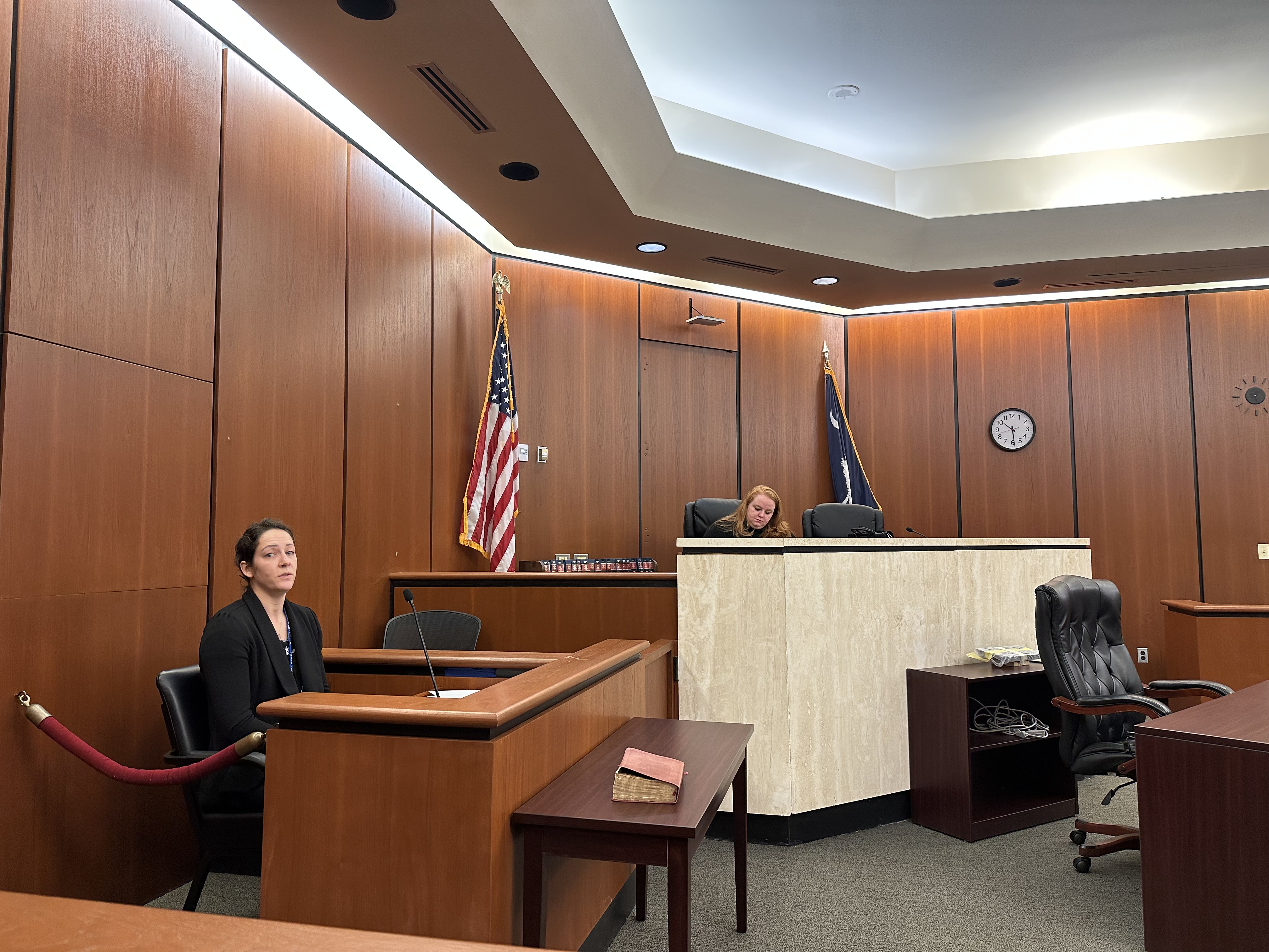 Image of a courtroom. A woman is sitting at the judge's bench, and a woman is sitting in the witness box.