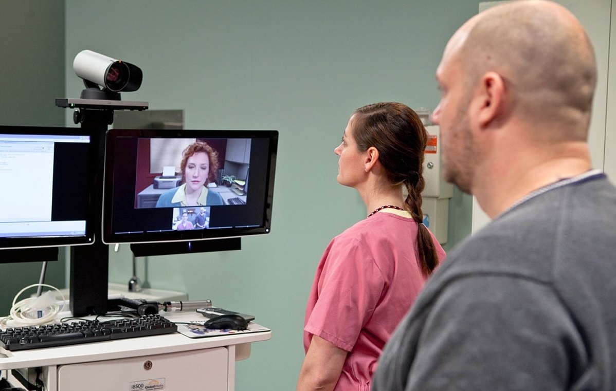 A telepsychiatry session in progress; a medical provider stands in front of a display showing a video call with a patient